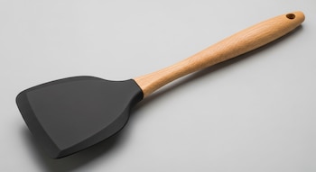 Espátula de cocina moderna con pala ancha de silicona negra mate y mango largo de madera clara, vista en ángulo sobre un fondo liso de tono gris.