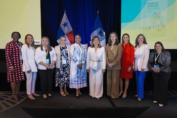 Nueve mujeres de pie con credenciales, frente a la bandera de Panamá y otra, una cortina azul y una pantalla con texto y el logo del BID