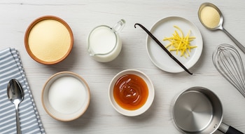 En una mesa de madera clara, se ven cuencos con sémola, azúcar, caramelo, una jarra con leche, una vaina de vainilla, ralladura de limón, una cuchara, un batidor y una cacerola.