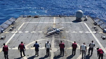 Dron gris en la cubierta de un buque de la Marina de EE.UU. con marineros alrededor y el océano azul visible al fondo