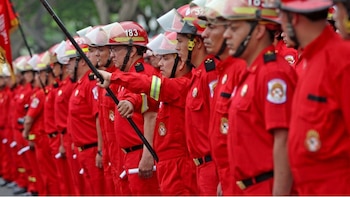 Una línea de bomberos en uniforme rojo con cascos protectores, algunos numerados. Un bombero sostiene un mástil. Fondo verde borroso