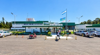 Edificio industrial con el logotipo de Granja Tres Arroyos, banderas en mástiles, personas caminando, una motocicleta roja y varios automóviles