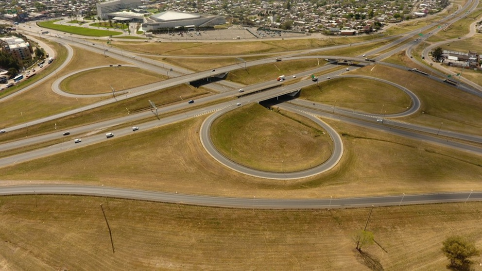 Vista aérea de un complejo nudo vial con múltiples carriles y puentes elevados, rodeado de terrenos secos y vegetación, con edificios urbanos al fondo