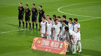 Jugadores de fútbol vestidos de blanco posan tras un cartel de España y México junto a cuatro árbitros en una cancha.