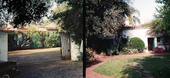 Vista de la fachada de una casa de una planta con paredes blancas y techo de tejas, rodeada de vegetación, con una entrada adoquinada y una puerta de metal