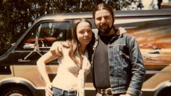 Un hombre barbudo y una mujer posan sonrientes frente a una furgoneta con franjas claras y un mural de un barco en un atardecer