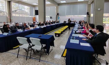 Varias personas sentadas alrededor de mesas azules en forma de U levantan la mano en una sala de reuniones