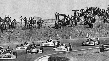 Imagen monocromática de una carrera de coches de Fórmula 2, con cuatro vehículos en la pista y una multitud de espectadores en la colina adyacente