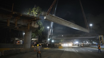 Fotografía nocturna de una grúa elevando una viga de hormigón sobre una autopista, con trabajadores y luces de la calle