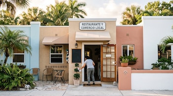 Fachada de un comercio pastel con el letrero Restaurante y Comercio Local y una persona junto a una planta en la entrada.