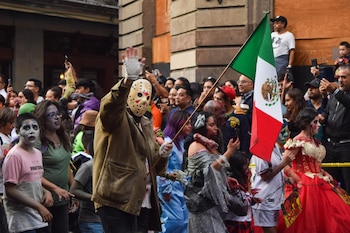 Una persona con máscara y la bandera de México saluda entre una multitud con disfraces y maquillaje de zombis en una calle urbana