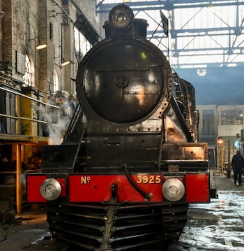 Frente de una locomotora a vapor negra con una placa frontal roja con el número 3925 dentro de un taller industrial
