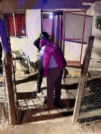 Un agente de uniforme negro y casco junto a una persona con chaqueta morada en la entrada de una casa iluminada de noche