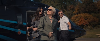 Meryl Streep, con gafas de sol y un blazer a rayas, saliendo de un helicóptero negro con franjas azules, acompañada por una mujer y un hombre