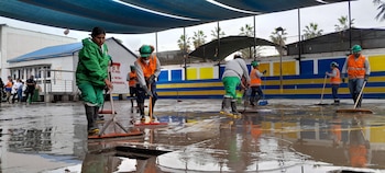Varias personas con chalecos naranjas y ropa de trabajo verde retiran agua de un patio escolar inundado con grandes escobas. Hay edificios, palmeras y una lona azul