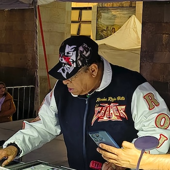 Un hombre con gorra, anteojos y chaqueta deportiva manipula una consola de sonido mientras una persona cercana sostiene un teléfono celular