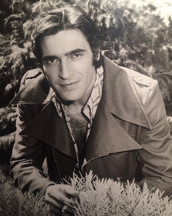 Retrato en blanco y negro de un hombre joven con chaqueta y camisa estampada, situado frente a un arbusto con un cigarrillo en la mano