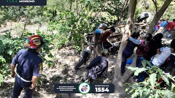 Un bombero y varias personas rodean un vehículo volcado en un área boscosa. Se observan árboles, vegetación y el logo de Bomberos Municipales Departamentales