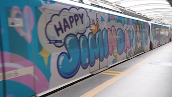 Vagón de tren azul y blanco con letras de colores que dicen Happy Jiminday y el retrato de un joven en el andén de una estación