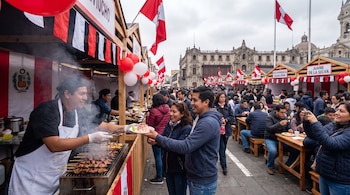 Una feria callejera con puestos de madera decorados con banderas peruanas y globos rojos y blancos. Un chef sirve anticuchos a una pareja. Personas comen y caminan.