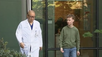 Un médico calvo con gafas y bata blanca camina junto a un joven de pelo rizado y suéter verde, frente a un edificio con ventanas