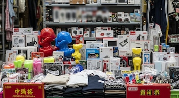 Puesto de mercado con una mesa llena de cajas, juguetes de plástico, ropa apilada, dispositivos electrónicos y utensilios domésticos. Se ven etiquetas en chino.