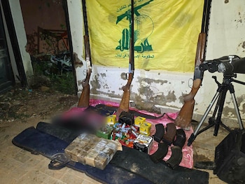 Armas incautadas sobre un suelo de tierra y hormigón, incluyendo tres escopetas, municiones, cargadores, un telescopio y una bandera amarilla de Hezbollah al fondo