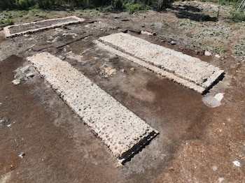 Vista aérea de un sitio arqueológico con dos estructuras rectangulares paralelas de piedra clara y una tercera más pequeña al fondo, rodeadas de tierra excavada y vegetación