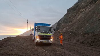 Un camión blanco transita por una vía de tierra junto a un cerro y el mar, al lado de un operario con casco y chaleco anaranjado