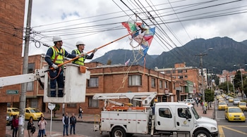 Dos hombres con uniforme de trabajo y casco en una cesta elevadora manipulan una cometa grande enredada en un tendido de cables aéreos.
