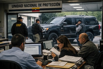 En una oficina de la UNP en Colombia, personas revisan documentos y computadores. Detrás, a través de la ventana, se observa a escoltas y un vehículo blindado oscuro.