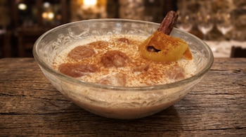 Cuenco de vidrio con leche, castañas cocidas, canela molida, una rama de canela y piel de cítrico sobre una tabla de madera.
