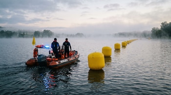 Dos rescatistas en una lancha inflable de rescate avanzan por un lago con niebla junto a una fila de boyas amarillas.
