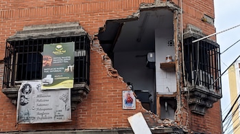 Edificio de ladrillo rojo con un gran agujero en la esquina. Dos ventanas con rejas de metal. Banners publicitarios colgando. Restos de escombros. Cables