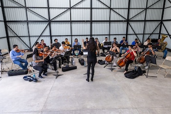 Una directora de espaldas lidera a una orquesta diversa de músicos tocando violines, chelos, trompetas, trombones y una tuba en un gran salón
