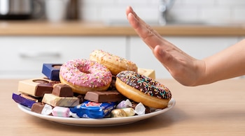 Una mano extendida sobre un plato blanco con donas de colores, chocolates variados y otros dulces, con una encimera de cocina de madera al fondo.