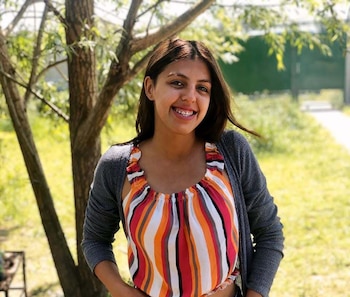 Una mujer joven con blusa a rayas de colores y suéter gris sonríe al aire libre junto a un árbol