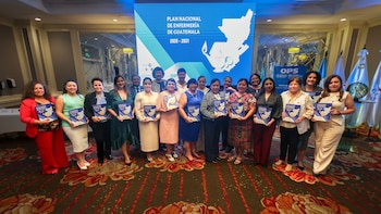 Grupo de profesionales de la salud, mayormente mujeres, posando con documentos del "Plan Nacional de Enfermería de Guatemala" frente a una pantalla