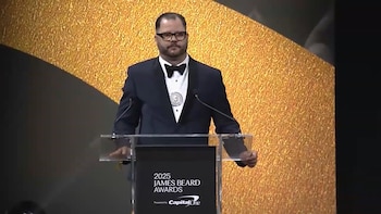 Hombre con gafas y barba, vestido de esmoquin, habla desde un podio transparente con el logo '2025 James Beard Awards'. El fondo es dorado y negro