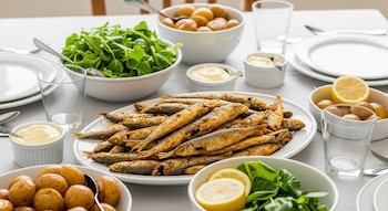 Mesa con mantel blanco, platos, vasos, cornalitos dorados, cuencos con papas, ensalada, gajos de limón, mayonesa y salsa.