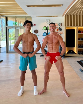 Cristiano Ronaldo y Cristiano Ronaldo Jr., ambos sin camiseta y en pantalones cortos, flexionan los bíceps mientras sonríen en un gimnasio moderno