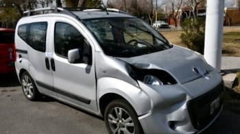 Primer plano de un furgón plateado con el parabrisas astillado y abolladuras en el capó y el parachoques delantero. Hay un coche rojo detrás