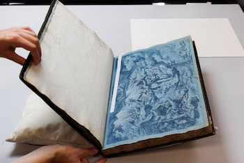Manos abriendo un libro antiguo de lomo oscuro sobre un cojín. La página izquierda tiene un bosquejo, la derecha muestra una ilustración monocromática azul