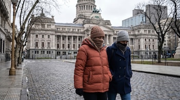 Dos personas, vestidas con abrigos gruesos y gorros, caminan por una calle adoquinada. Un gran edificio de piedra de estilo clásico se alza en el fondo bajo un cielo nublado.