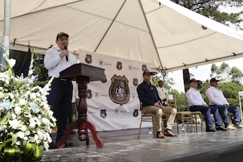 Un hombre de pie habla por micrófono en un podio, tres hombres sentados en sillas, toldo blanco, arreglo floral, bandera, letrero de Ministerio de Gobernación