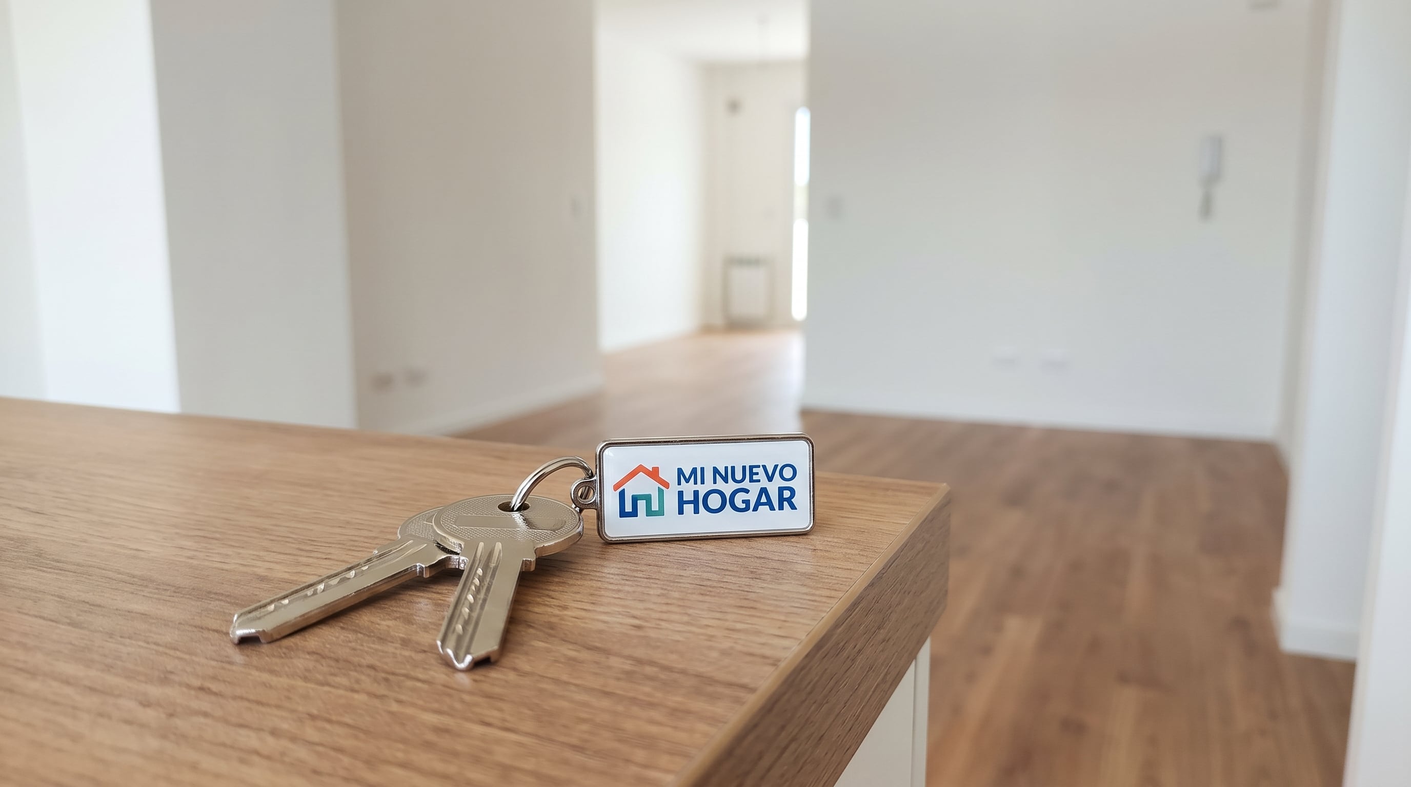 Llaves de una vivienda nueva reposan sobre una mesa de madera en un espacio interior vacío, con un llavero que dice