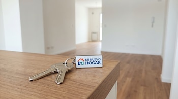 Dos llaves plateadas con un llavero rectangular blanco con el texto 'Mi Nuevo Hogar' sobre madera. Fondo de habitación vacía con paredes blancas.