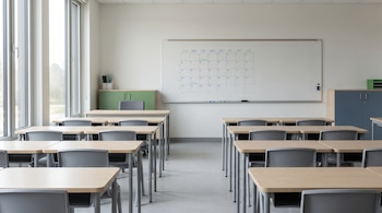 Aula moderna con paredes blancas, grandes ventanas, filas de escritorios de madera clara y sillas grises. Pizarrón blanco con calendario escolar y armarios al fondo.