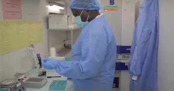 Un hombre con bata azul, mascarilla y guantes sostiene un tubo de ensayo junto a una gradilla con frascos en un laboratorio