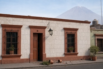 Fachada de piedra blanca con bordes terracota, una puerta de madera abierta, macetas rotas en el suelo y un volcán al fondo.